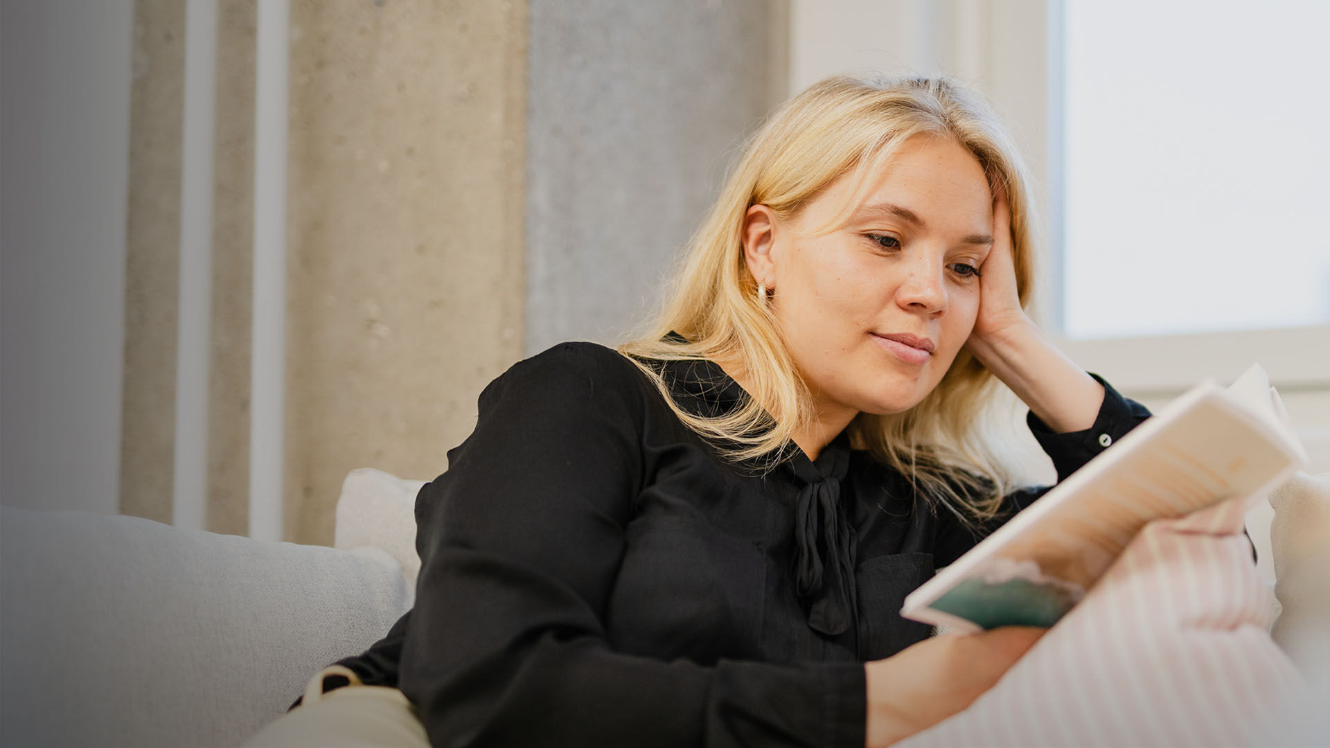 Nuori nainen lukee kirjaa  sohvalla tyynyyn nojaten
